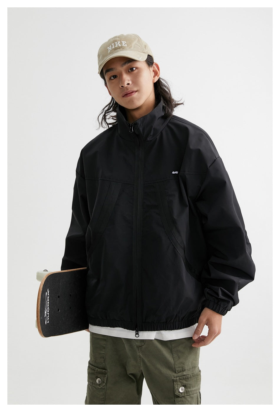 Model wearing a black zip-front jacket with a stand collar and curved front pockets, holding a skateboard.