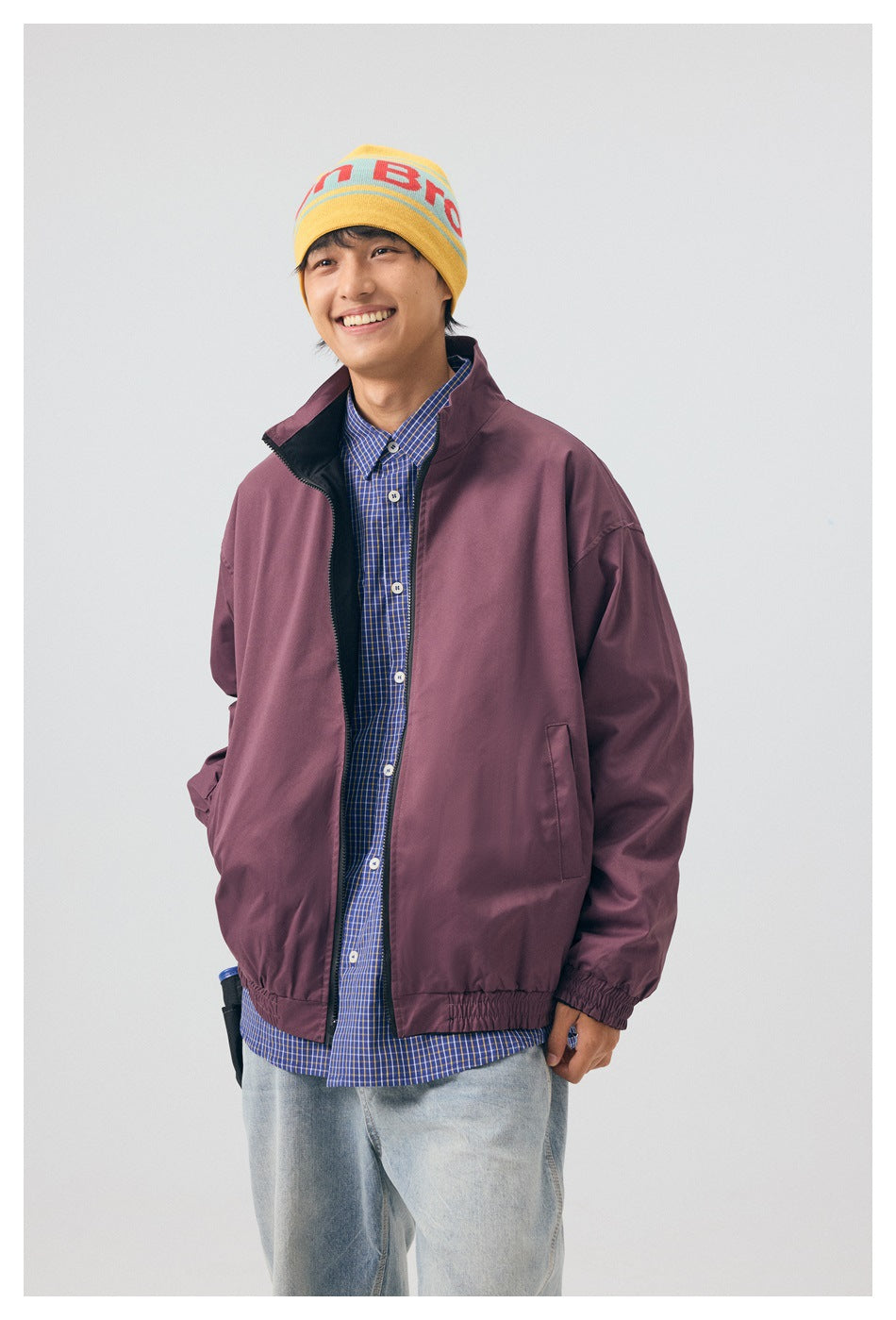Model wearing a plum zip jacket over a blue checked shirt, light jeans, and a yellow graphic beanie.