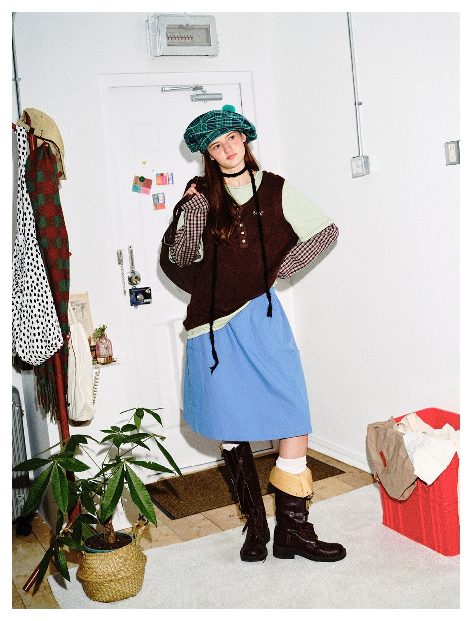 Model wearing a green plaid beret, layered burgundy and sage top, blue skirt, and dark boots, in a standing pose.