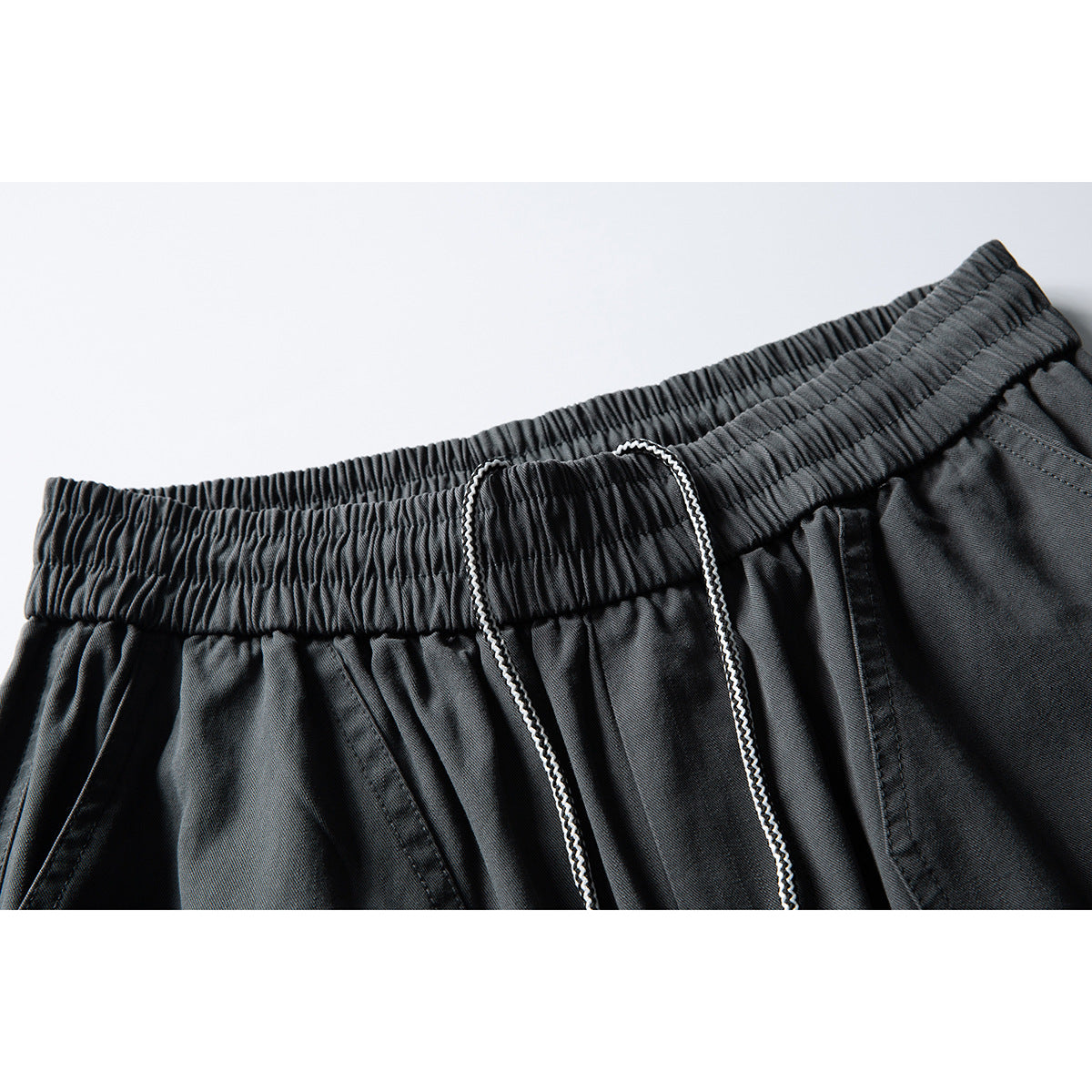 Close-up of charcoal pants showing the gathered elastic waistband, white drawcord, and front pocket seam.