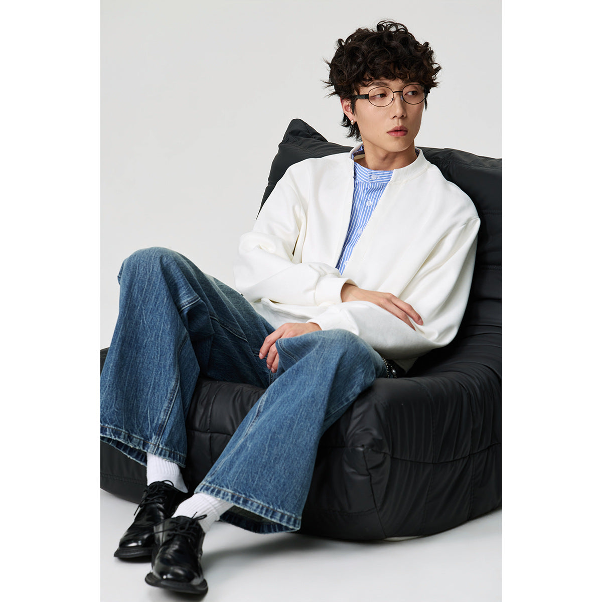 Seated model wearing a white split-neck pullover over a blue striped shirt with loose blue jeans.