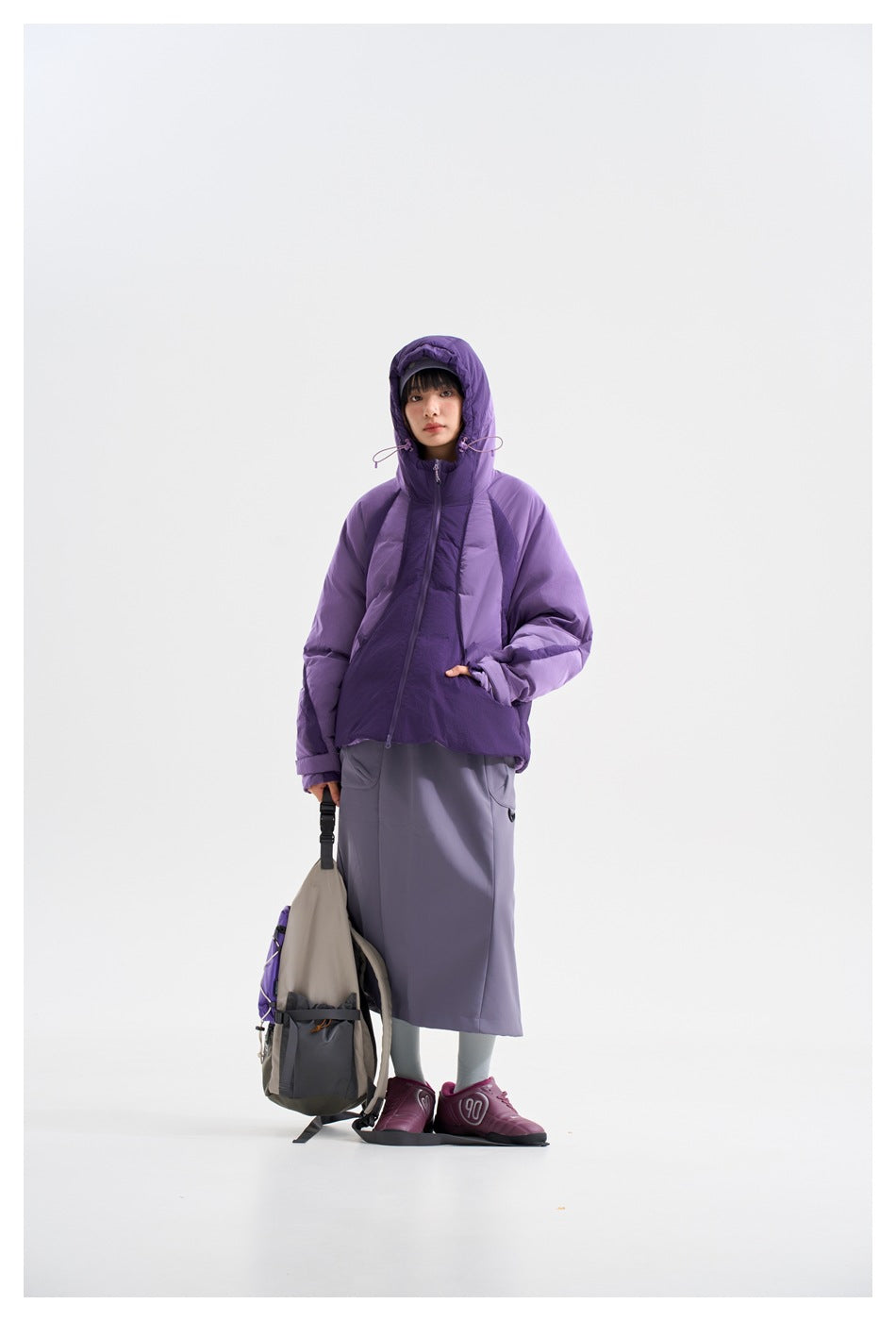 Model wearing a purple hooded jacket zipped to the neck, with a gray skirt and matching beanie.