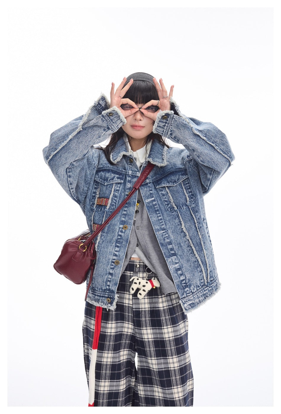 Model in a distressed blue denim jacket and navy plaid pants, posing with hands around her glasses.