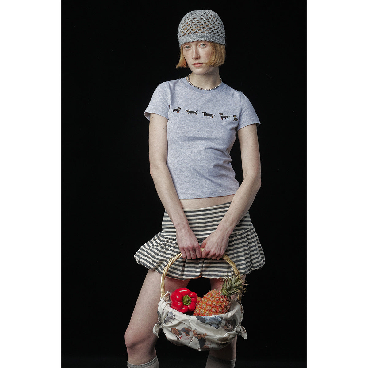 Model in a light gray dachshund-print T-shirt and striped skirt, holding a basket of fruit.