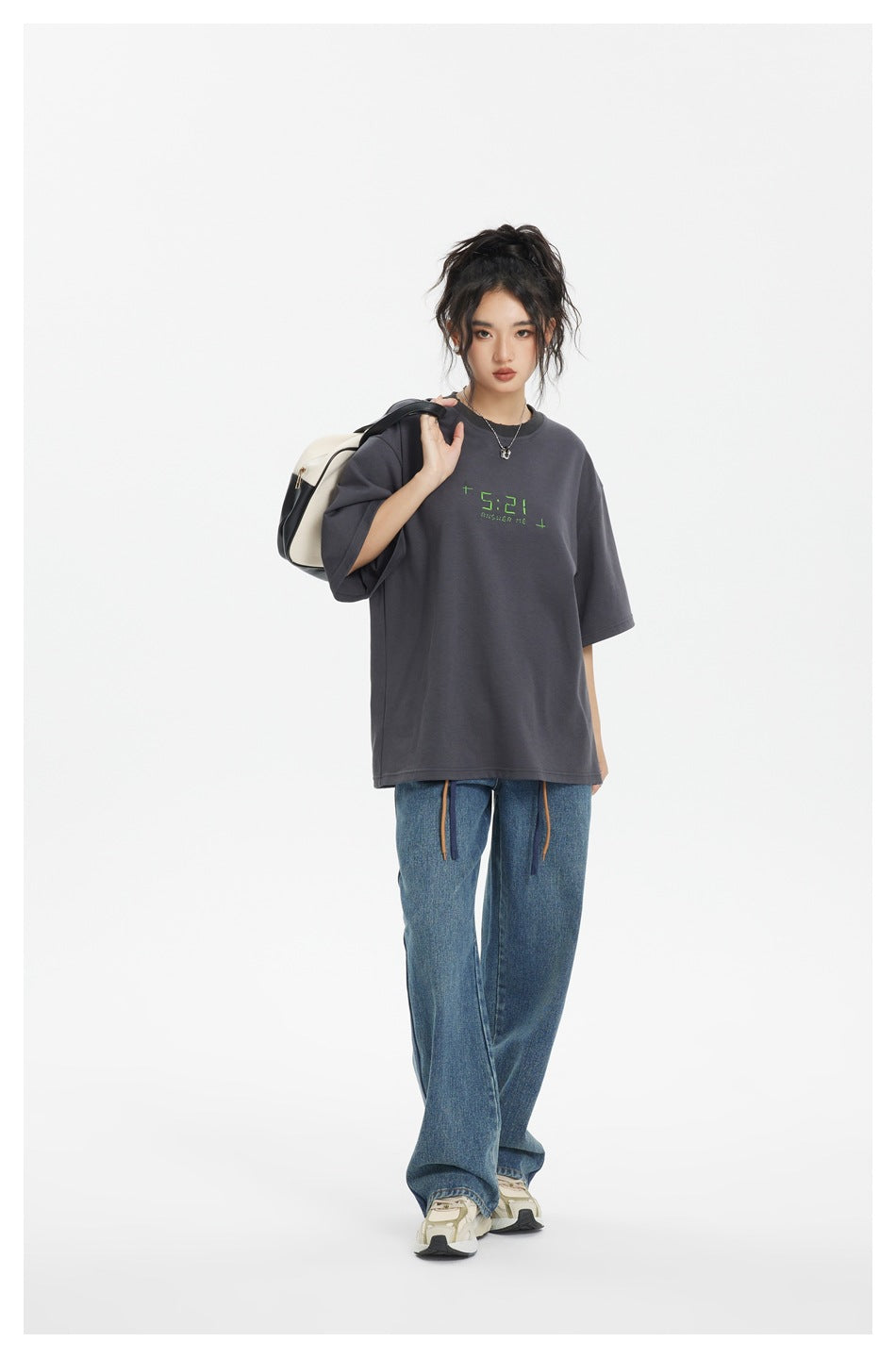 Model wearing an oversized charcoal T-shirt with green chest lettering and loose blue jeans.
