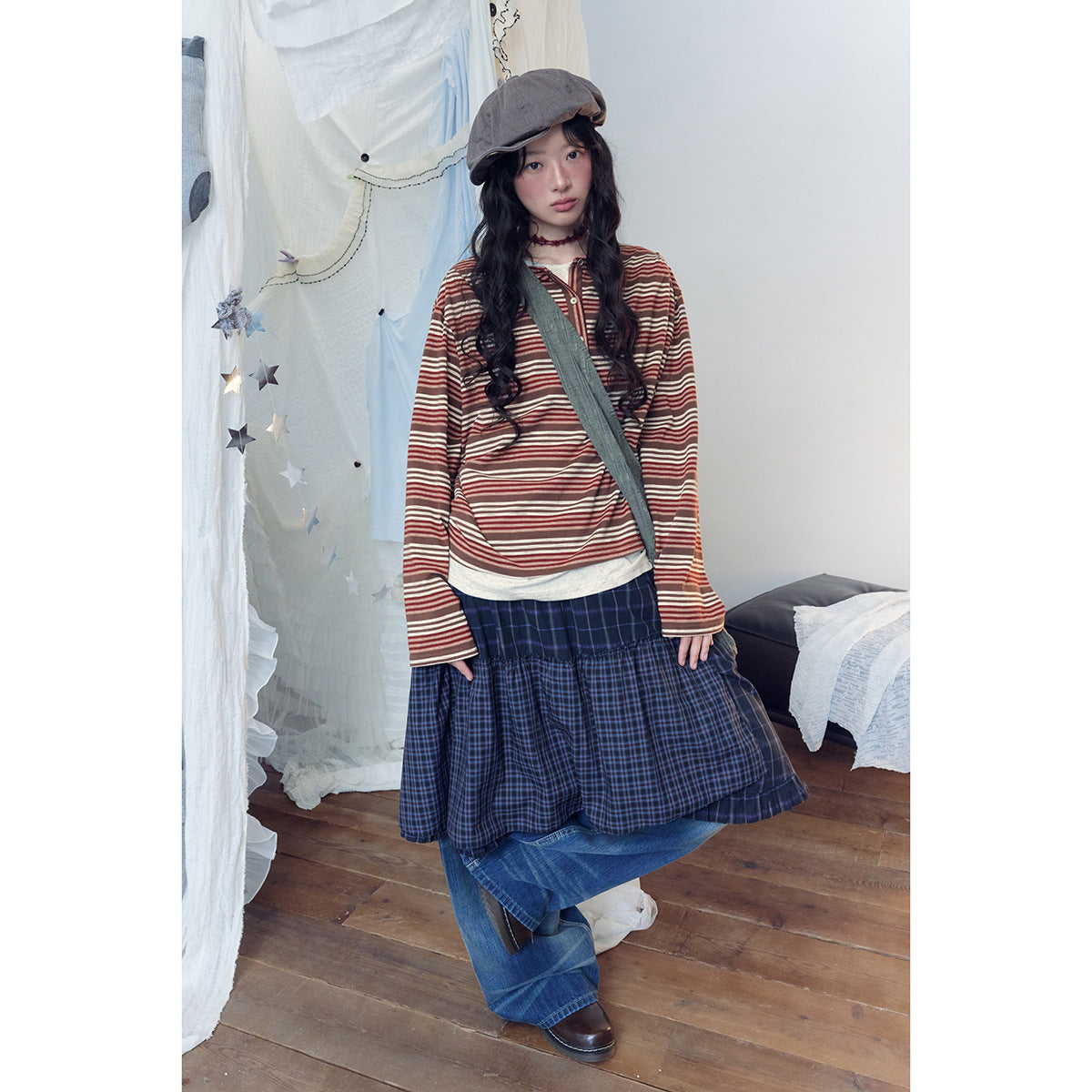 Model wearing a rust-and-cream striped henley, navy plaid layered skirt, wide-leg jeans and a gray cap.