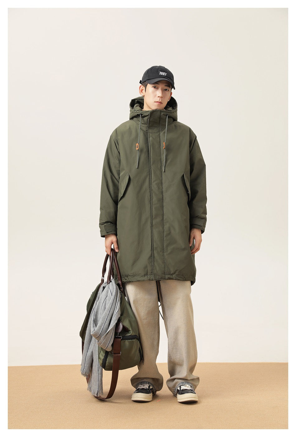 Full-length front view of a model wearing a dark olive hooded parka with orange cord toggles.