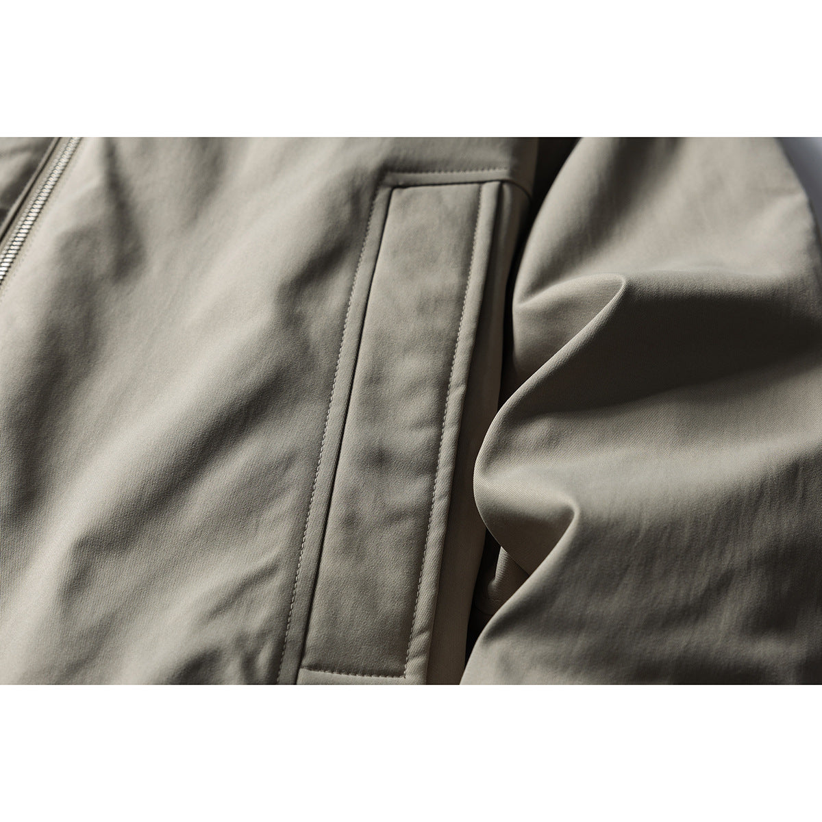 Close-up of a gray-beige jacket's slanted welt pocket, front zipper, and sleeve.