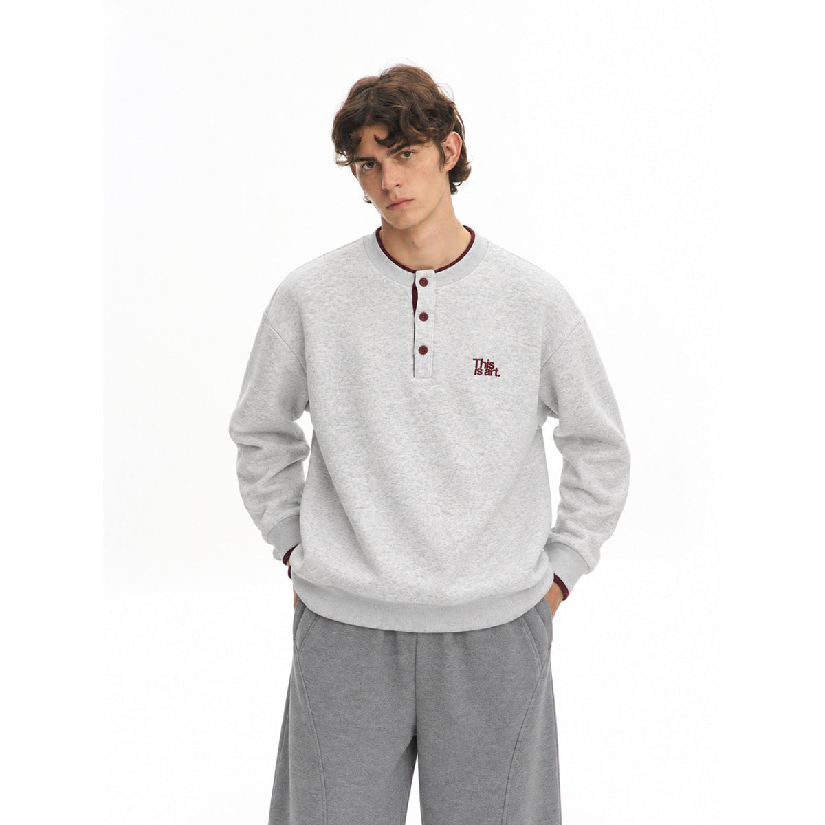 Model wearing a light gray marled Henley sweatshirt with burgundy buttons and chest lettering, paired with gray trousers.