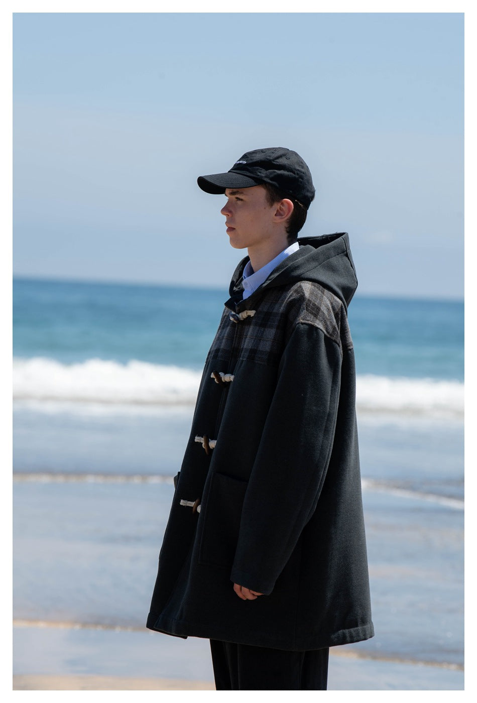 Front view of a model wearing a charcoal hooded coat with a plaid yoke, rope-and-toggle closures, and patch pockets.