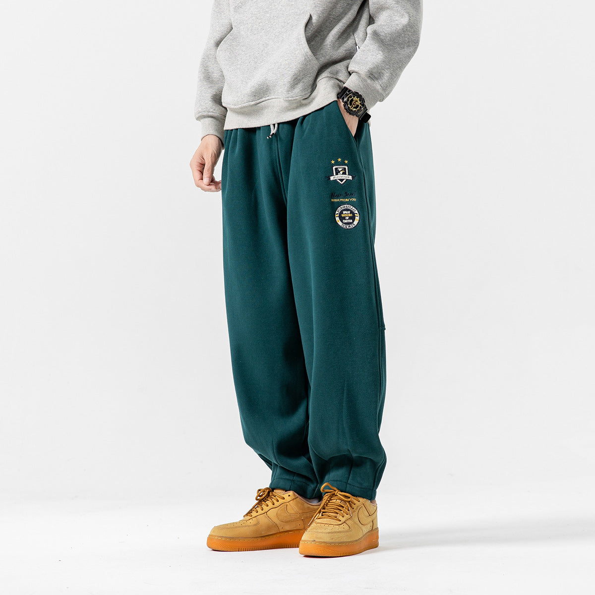 Model wearing deep teal relaxed joggers with a drawstring waist and embroidered thigh patches.