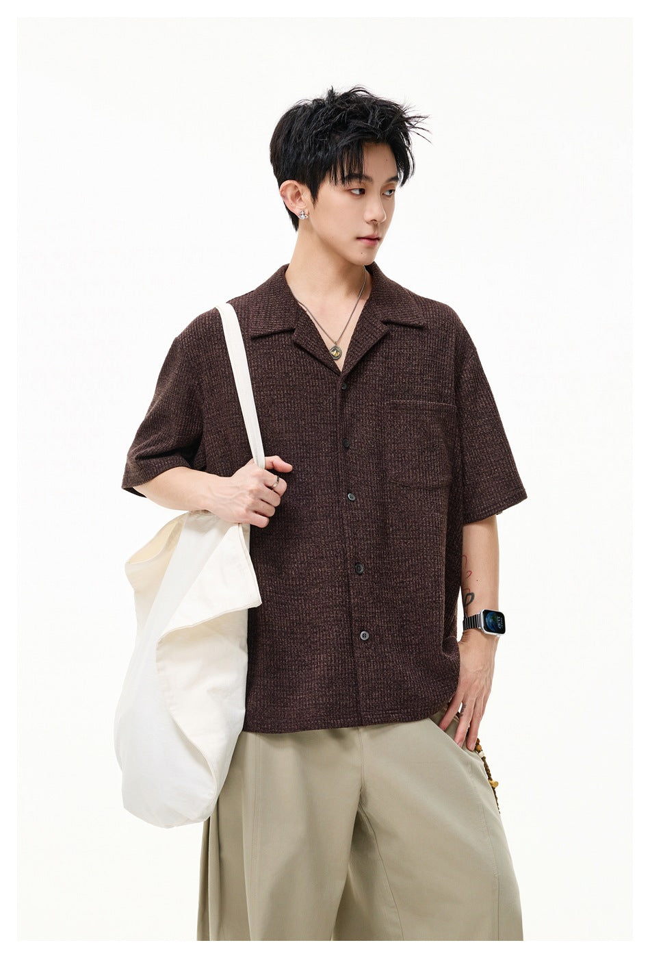 Model in a brown short-sleeve button-up shirt with a notched collar and chest pocket, carrying a canvas tote.