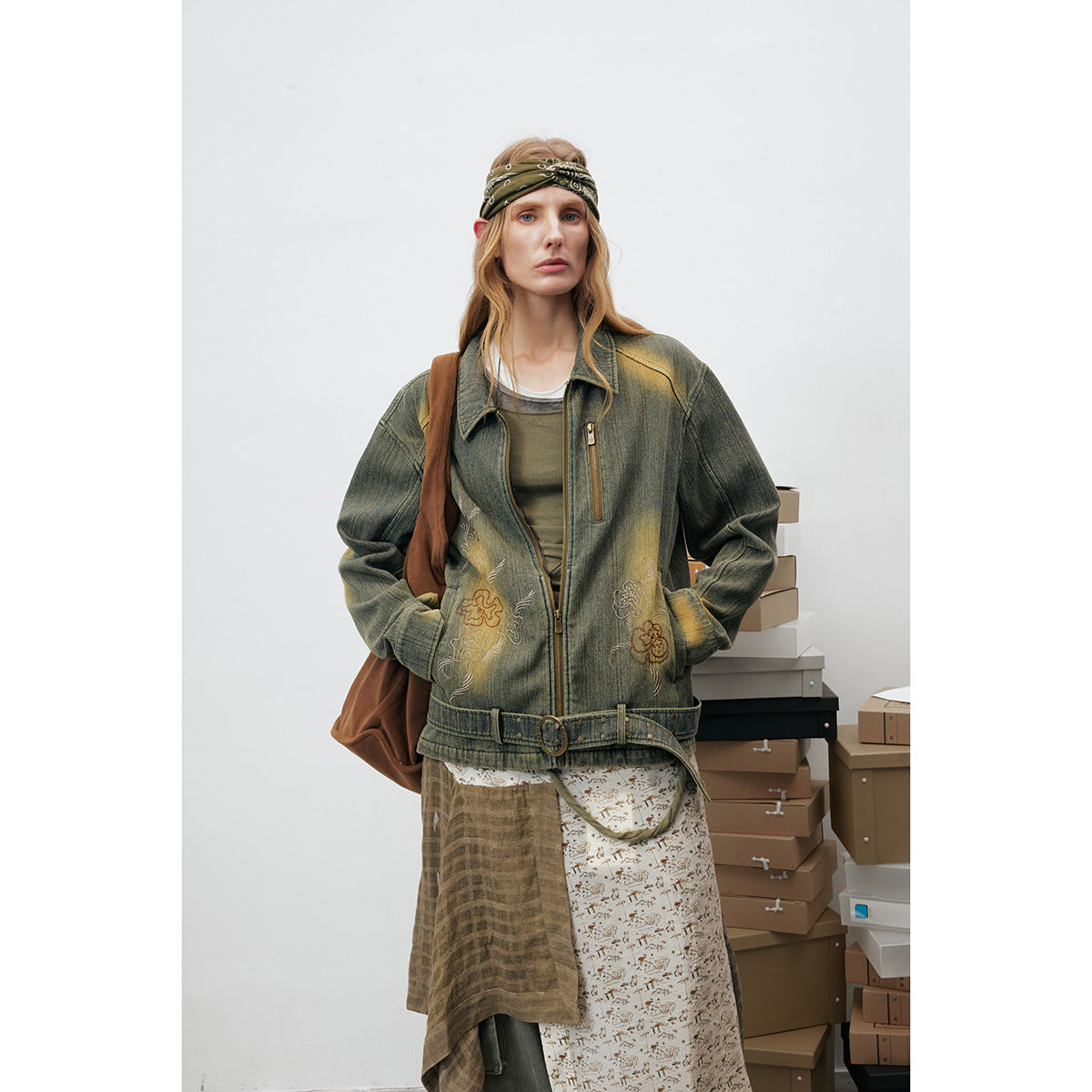 Model wearing an olive distressed jacket with floral embroidery, a belt and layered patterned garments.