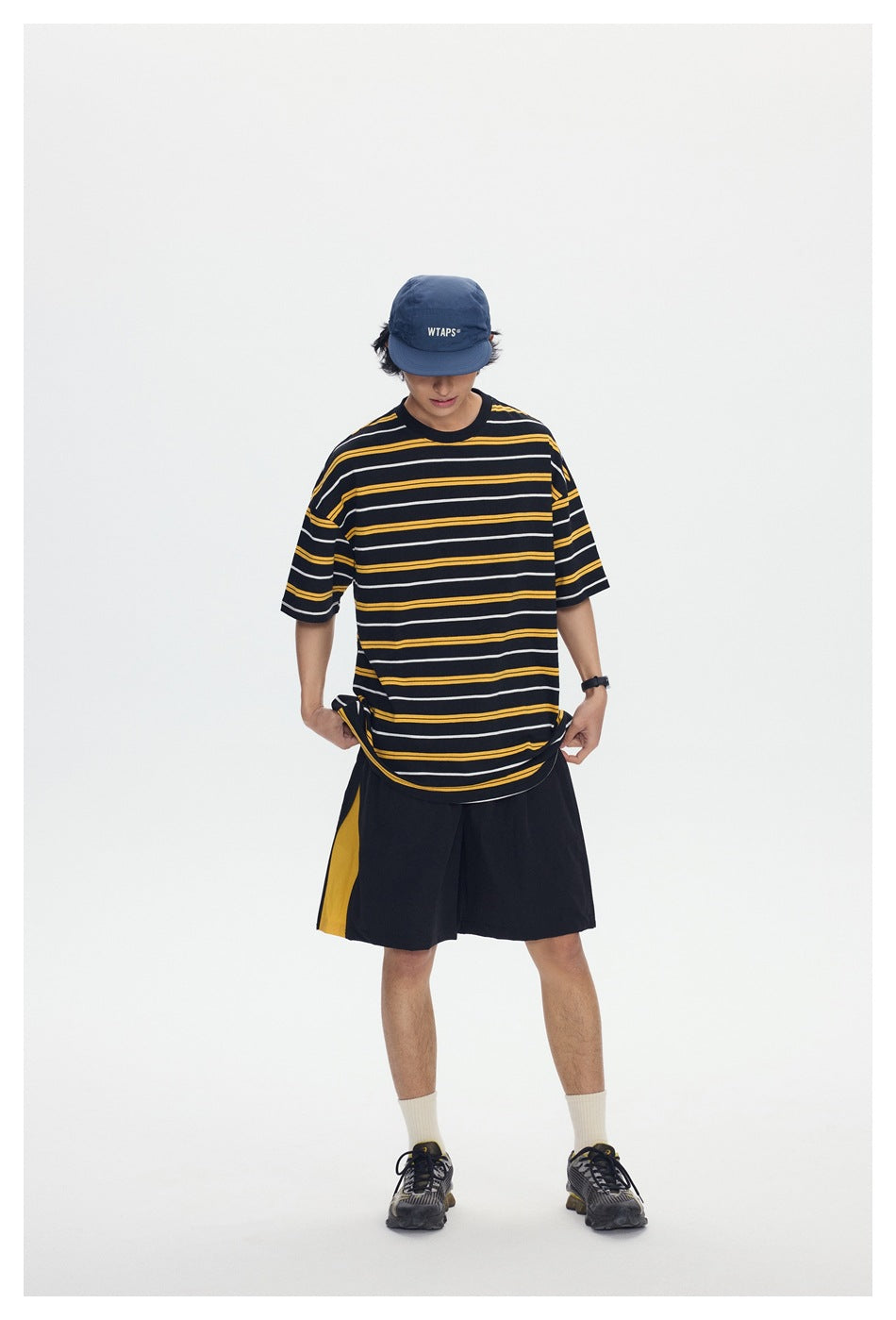 Front view of a model wearing a black T-shirt with yellow and white horizontal stripes and loose black shorts with a yellow
