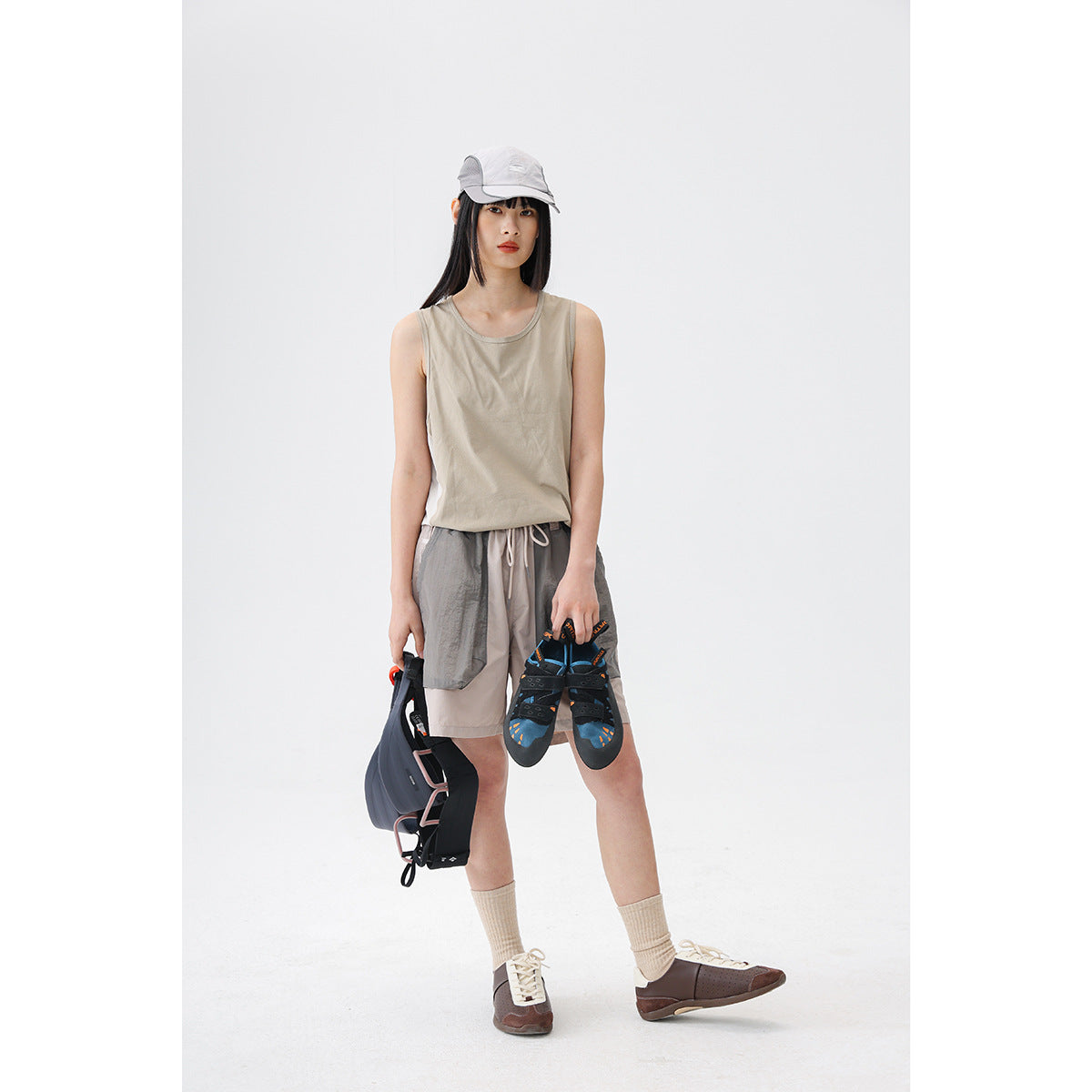 Model wearing a light beige sleeveless tank and two-tone gray drawstring shorts, holding outdoor gear.