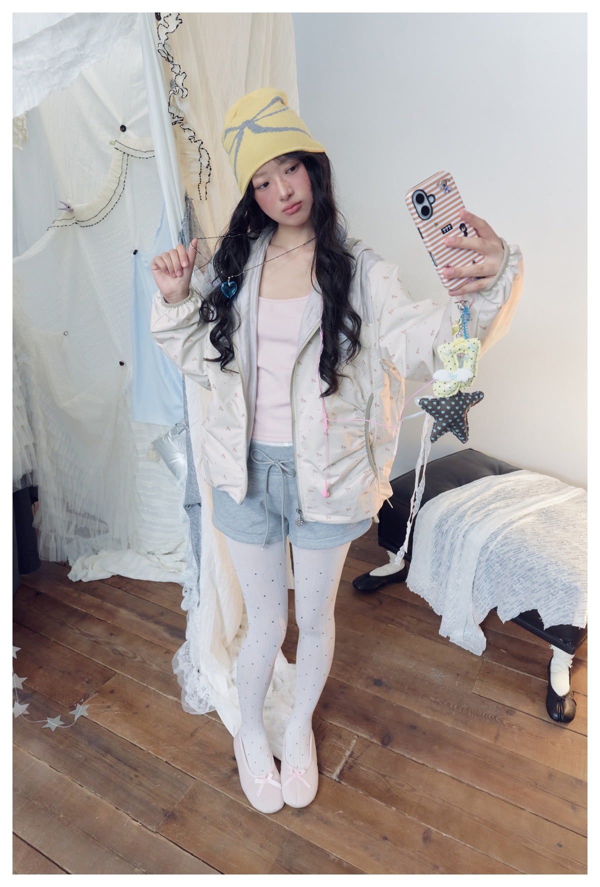 Model posing in a pale floral zip jacket, gray shorts and white dotted tights, wearing a yellow bow-pattern beanie.