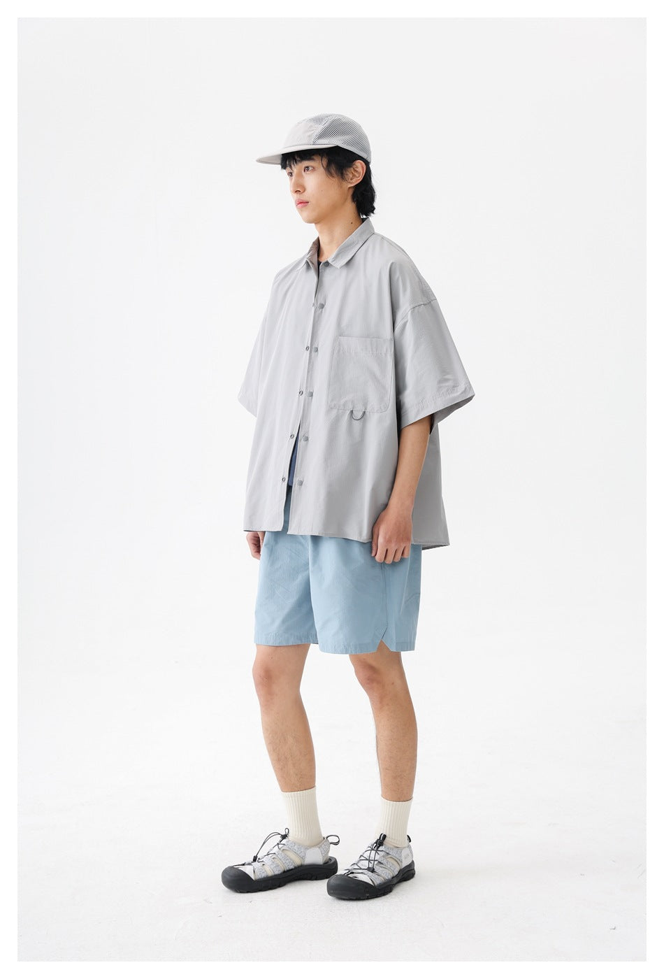 Model wearing a light gray oversized short-sleeve button-up shirt with a chest pocket, shown from the front at an angle.