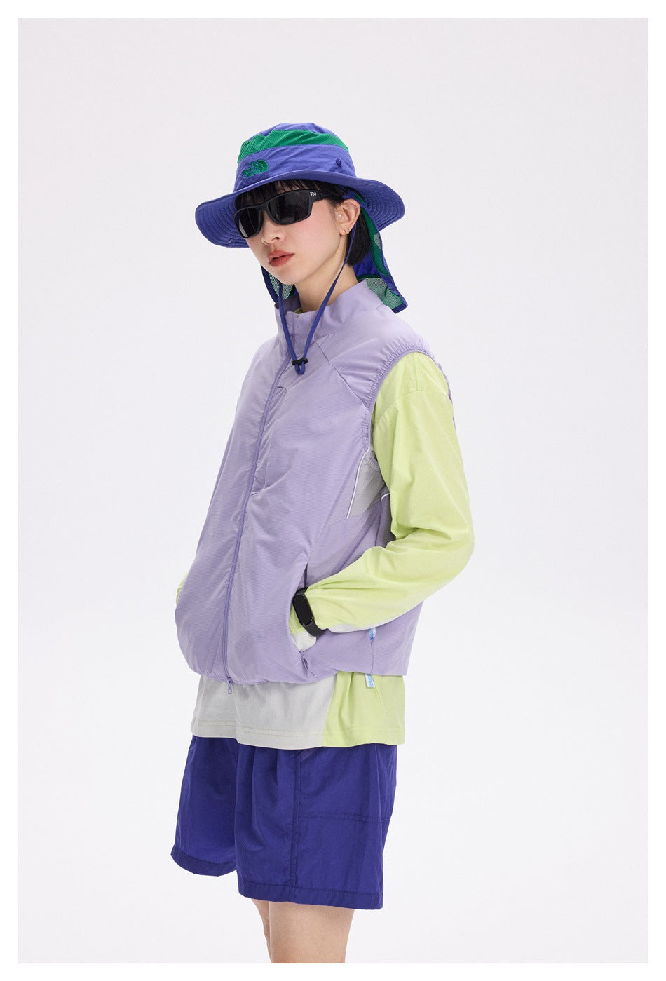 Close view of a model wearing a lavender zip vest over a lime long-sleeve top, blue shorts, and a wide-brim sun hat.