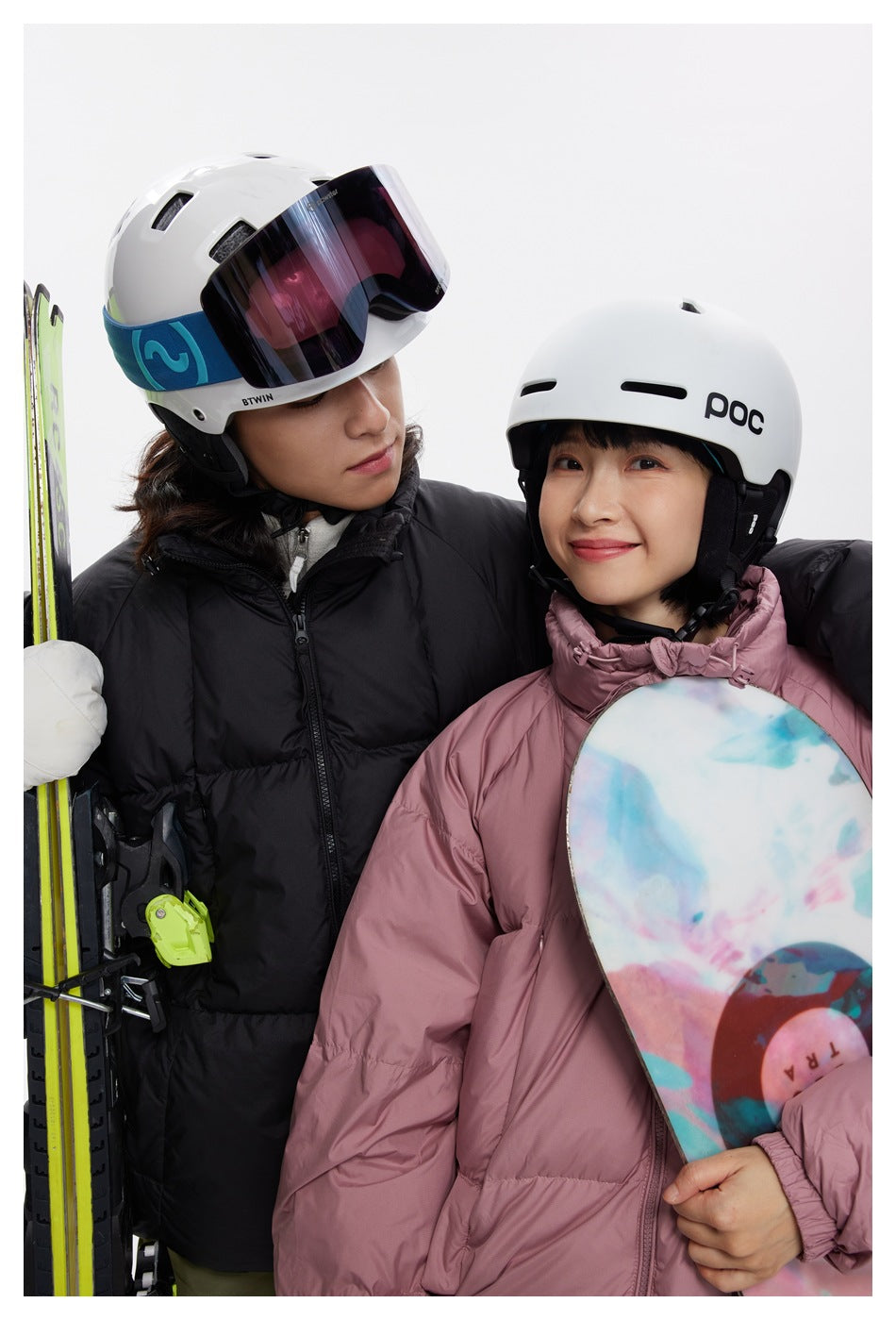 Close view of two models wearing black and dusty pink puffer jackets with ski helmets and a snowboard.