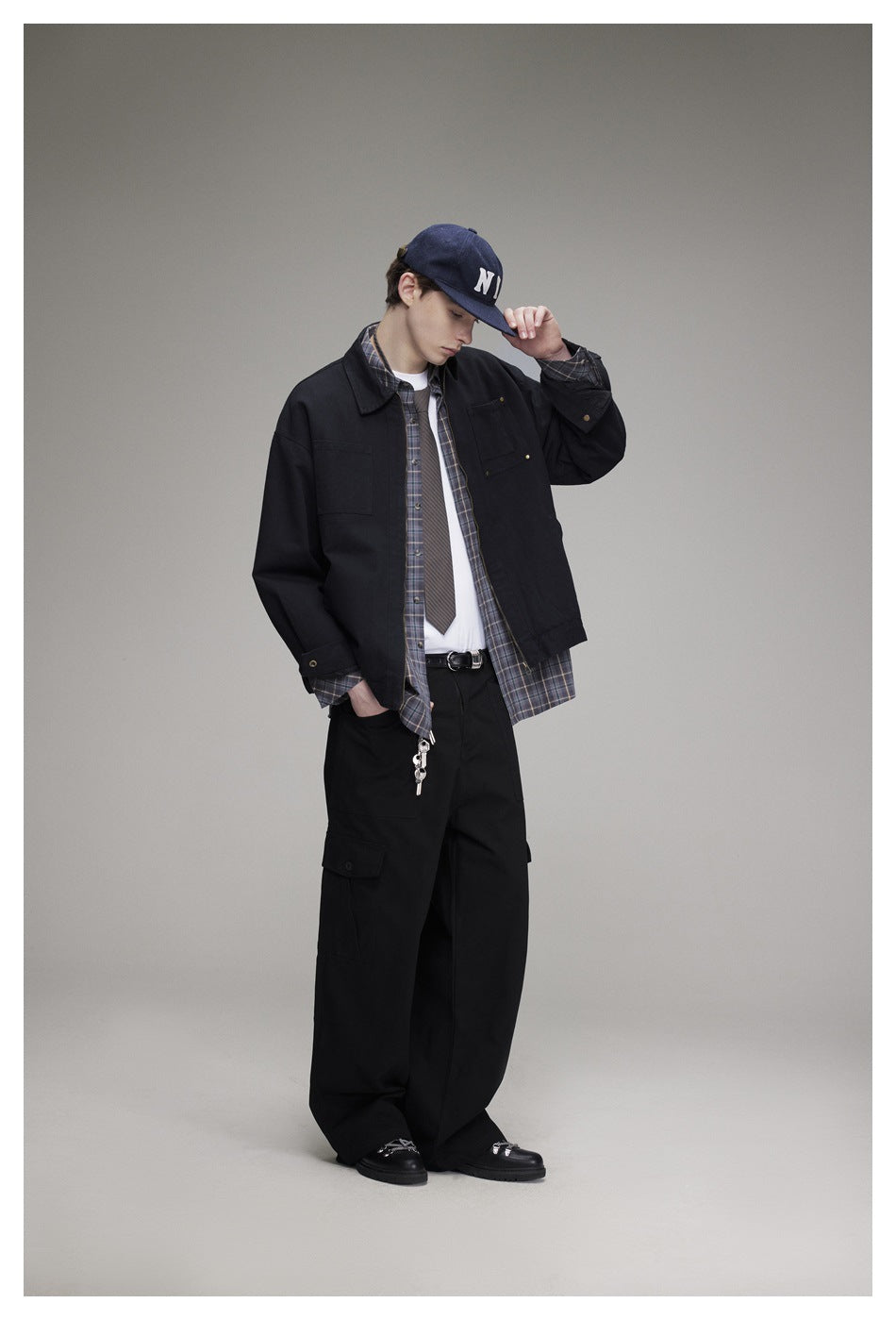 Model wearing a black zip-front jacket over a plaid shirt and white tee, with loose black cargo pants.