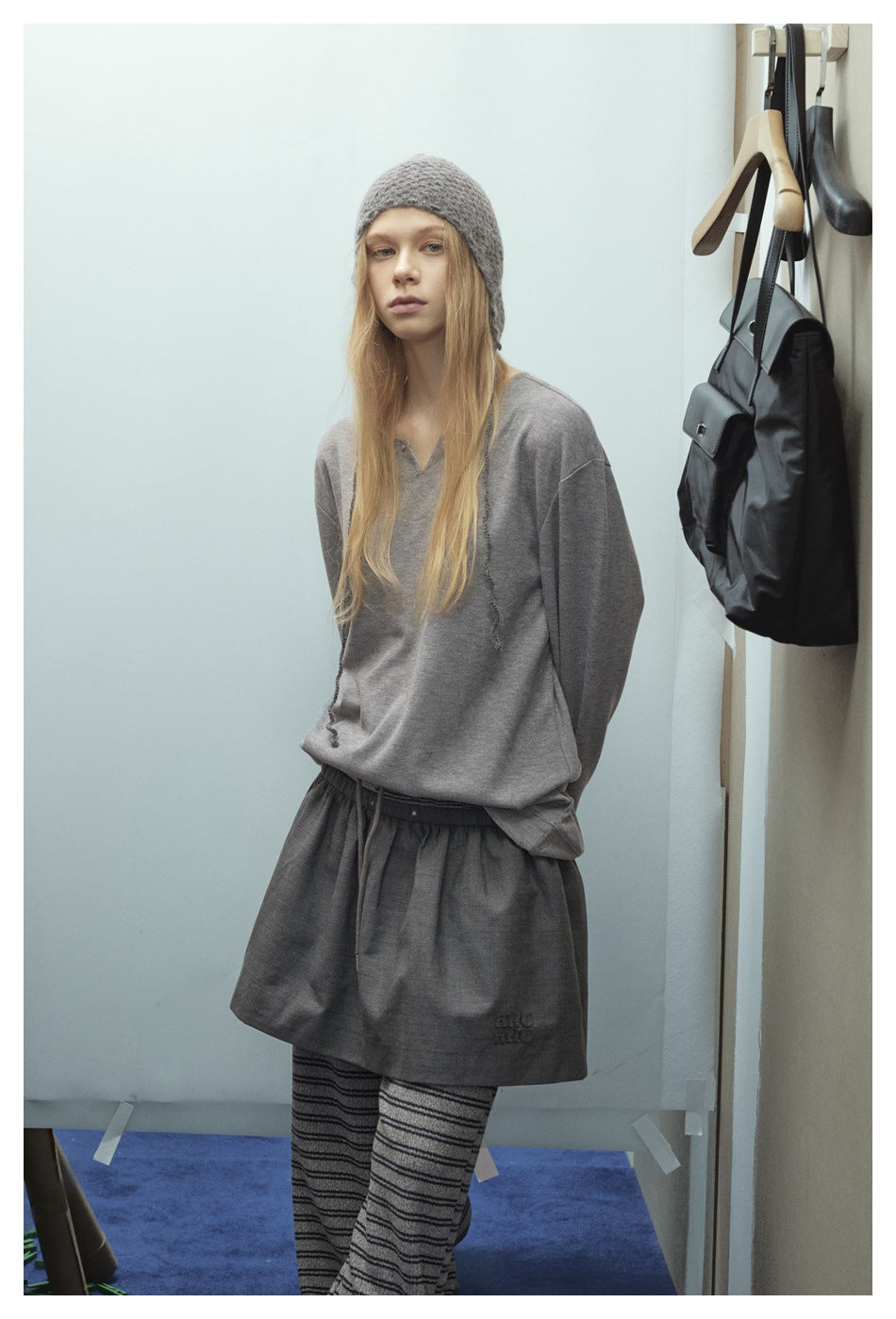 Model wearing a gray pullover and drawstring shorts over striped trousers, with a gray knit hat.