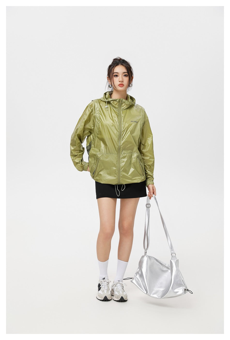 Model wearing an olive green hooded zip jacket over a black mini skirt, holding a silver bag, full-length view