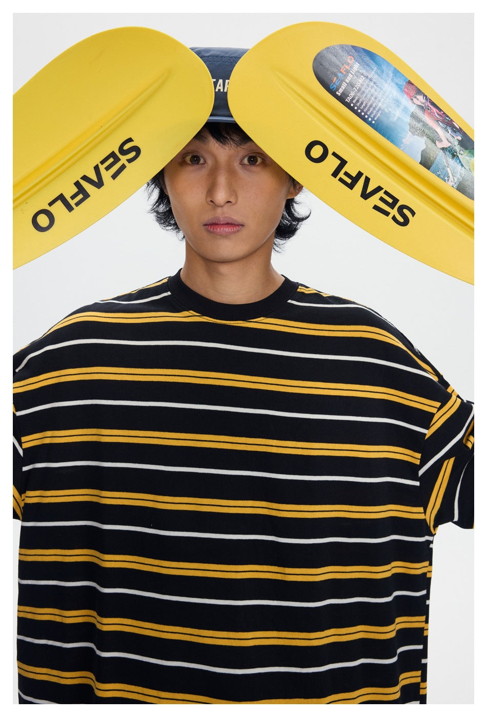 Close view of a model wearing a black oversized T-shirt with yellow and white horizontal stripes; yellow paddles frame his