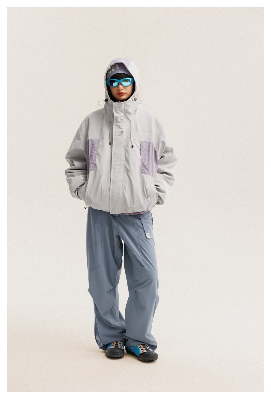 Model wearing a light gray hooded jacket with lavender panels and loose slate-blue pants, shown full length from the front.