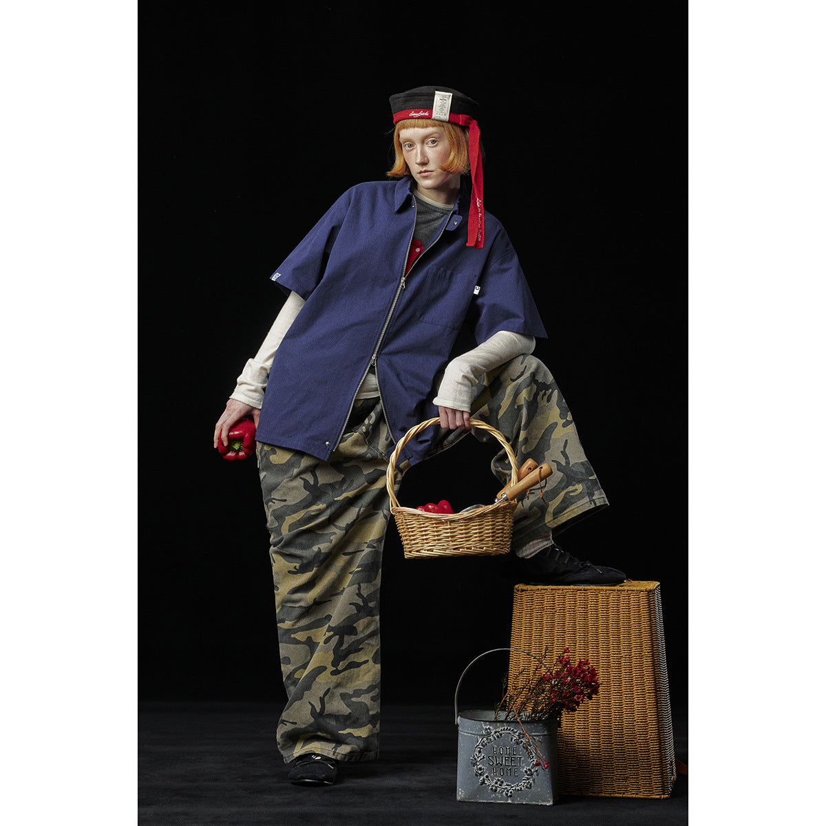 Model wearing an oversized navy short-sleeve zip shirt over layered tops and loose camouflage-print trousers.