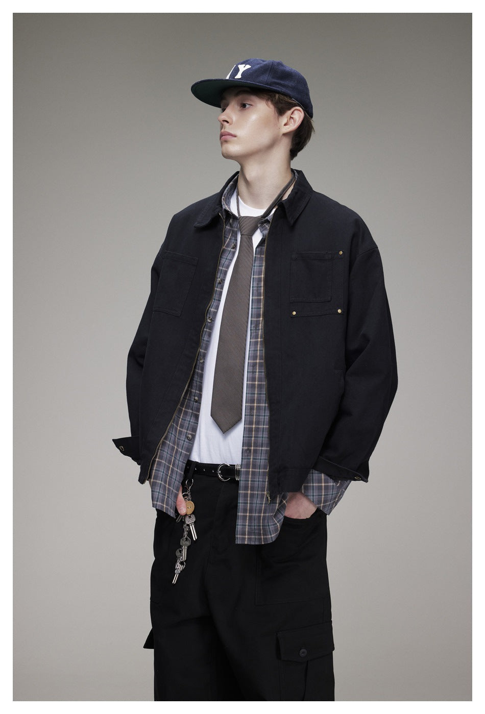 Model wearing a black zip-front jacket over a plaid shirt and white tee, with black cargo pants.