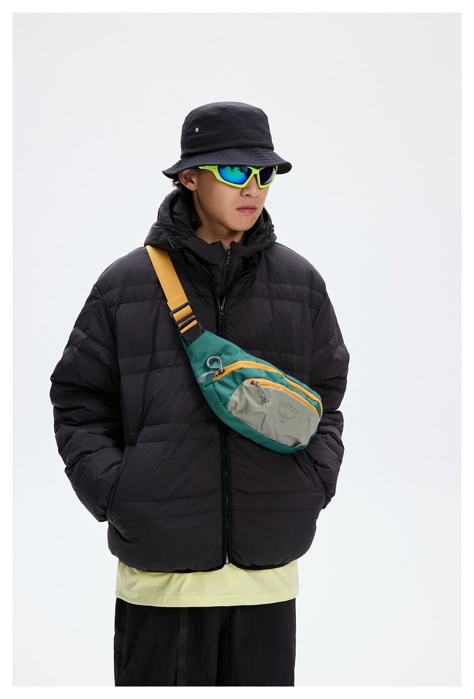 Model wearing a black hooded puffer jacket with a teal and gray crossbody bag and a yellow strap.