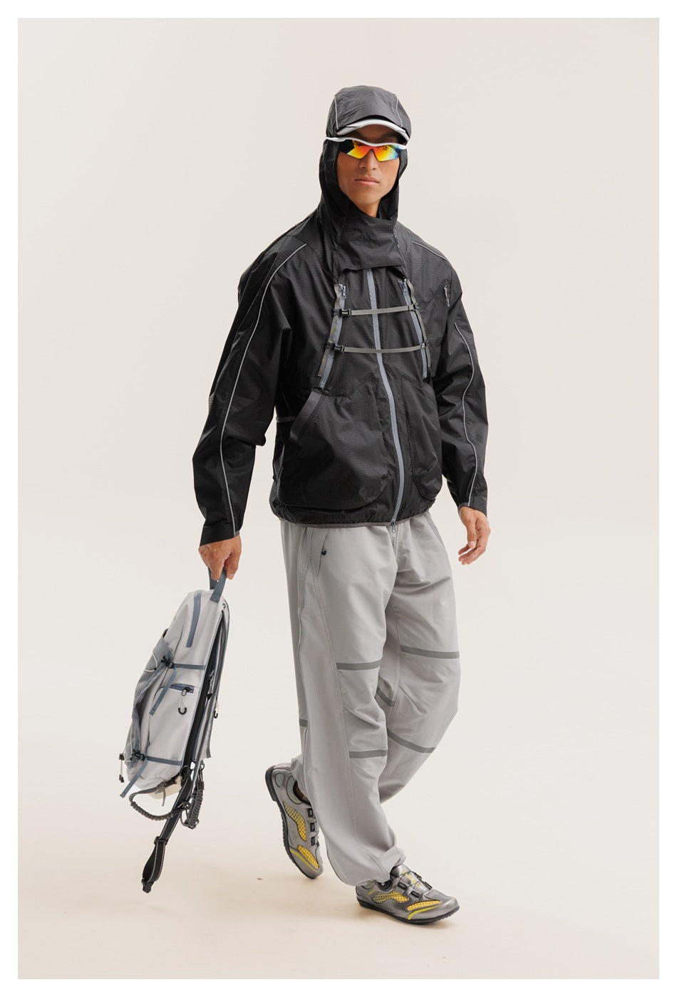 Model wearing a black hooded jacket with chest straps and gray pants, carrying a light gray backpack.