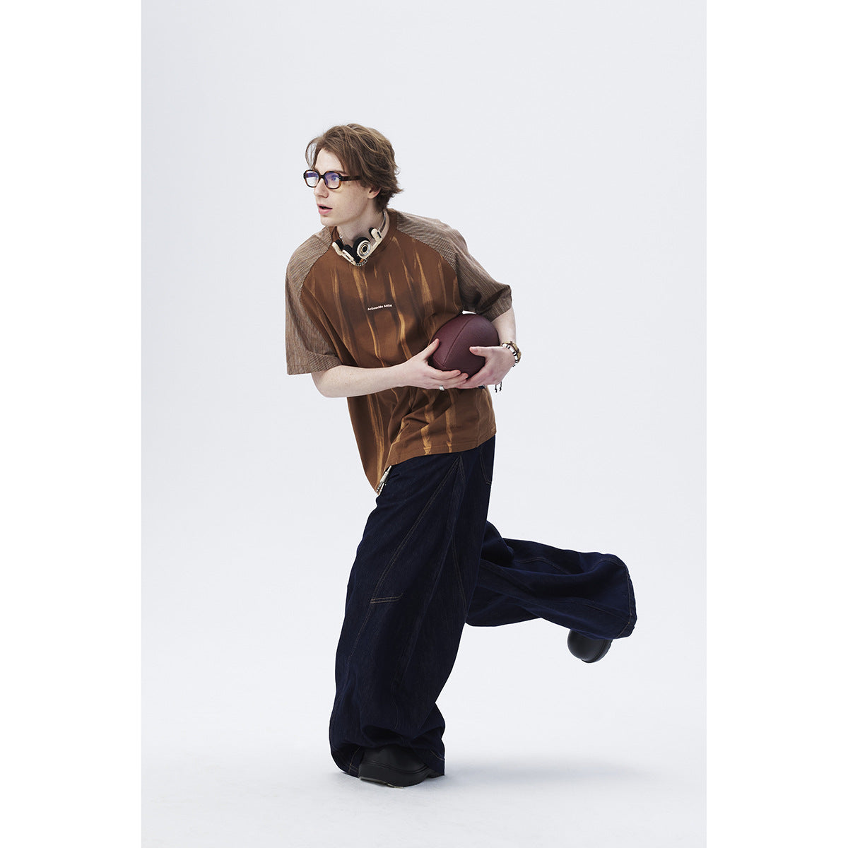 Model wearing a brown striped shirt with gray patterned sleeves and wide dark jeans, holding a ball.