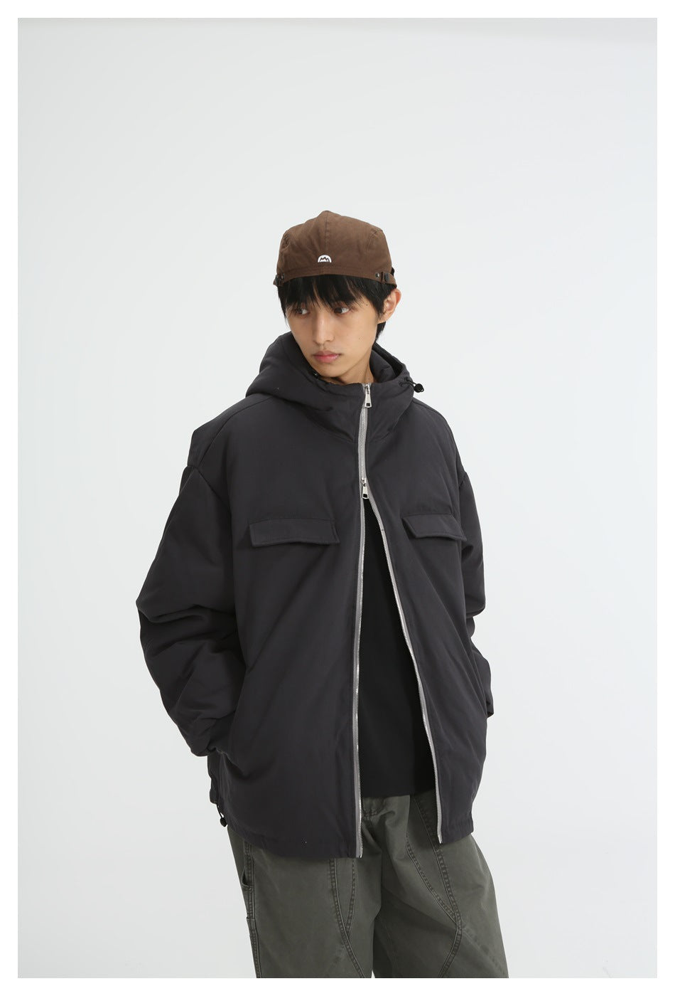 Model wearing black hooded padded jacket, unzipped front, with brown cap and olive green pants