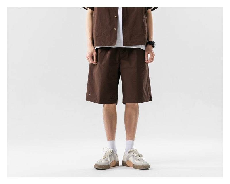 Cropped model facing forward in a dark brown short-sleeve button shirt and matching knee-length shorts.