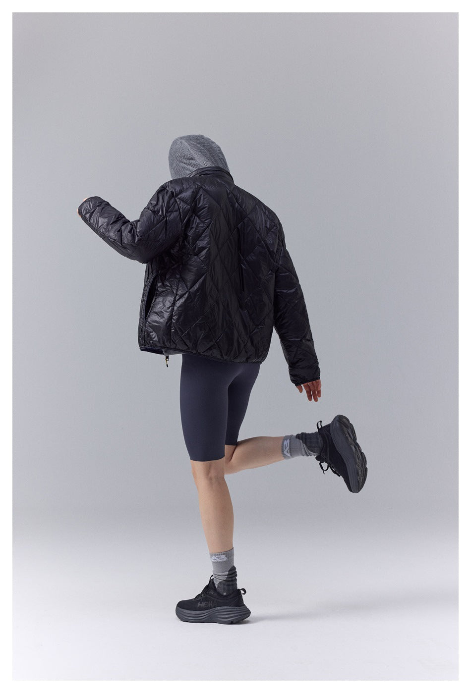 Model seen from the back in black diamond-quilted puffer jacket, gray knit hood and dark shorts