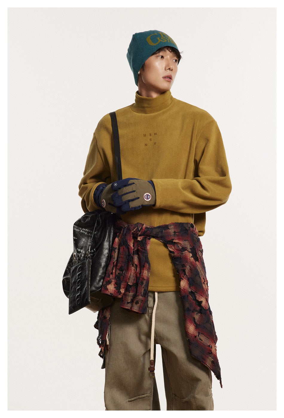 Model wearing a mustard high-neck pullover and teal beanie, with gloves, a black shoulder bag, and a plaid shirt tied at the