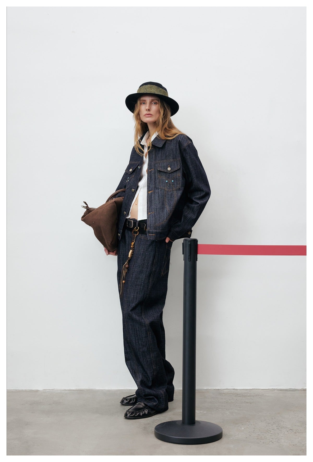 Model leaning on a stanchion wearing a dark denim jacket over a white shirt with matching wide-leg denim pants
