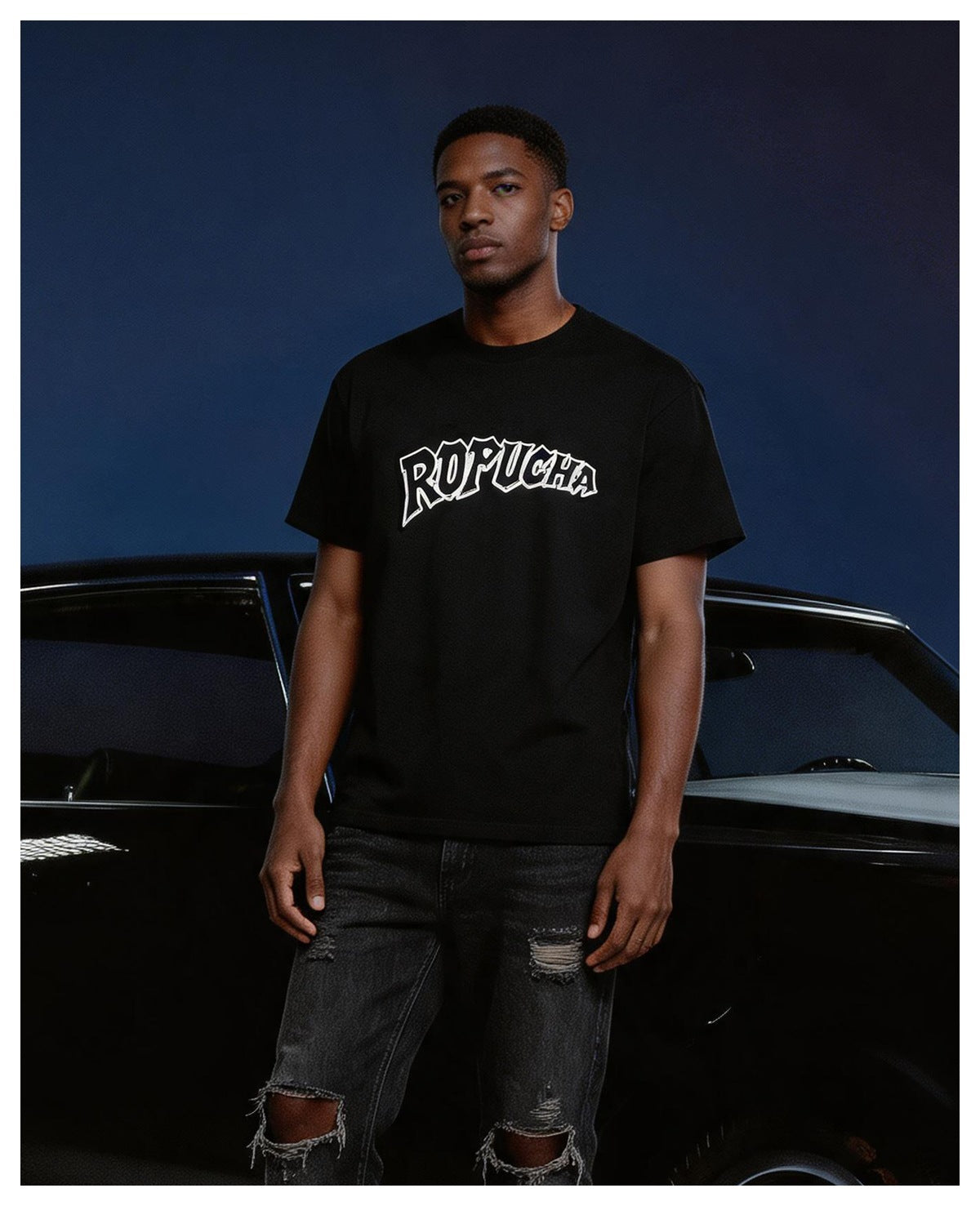 Man wearing a black ROPUCHA graphic T-shirt and ripped dark jeans standing in front of a black car against a blue backdrop