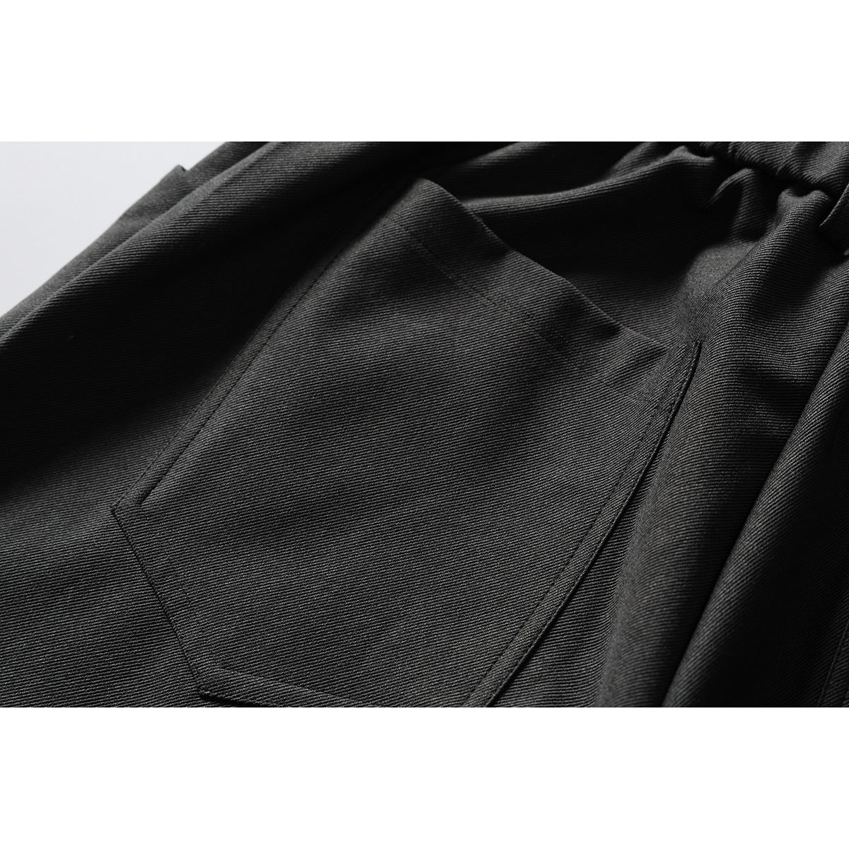 Close-up of a stitched patch pocket on dark charcoal trousers, with the gathered waistband visible above.