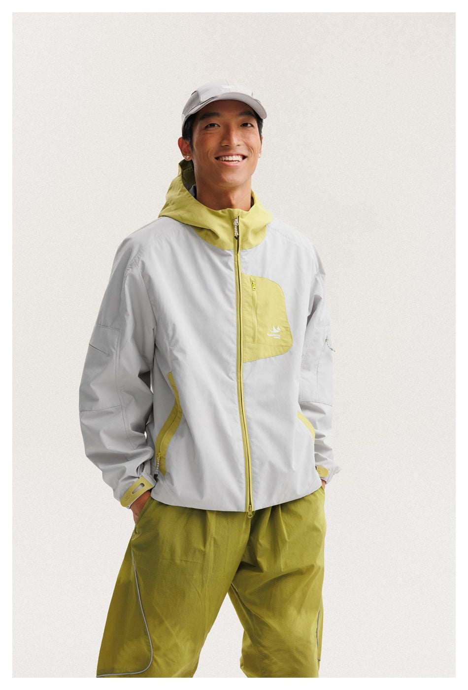 Mid-length front view of a smiling model in a light gray jacket with yellow-green hood and pocket accents.