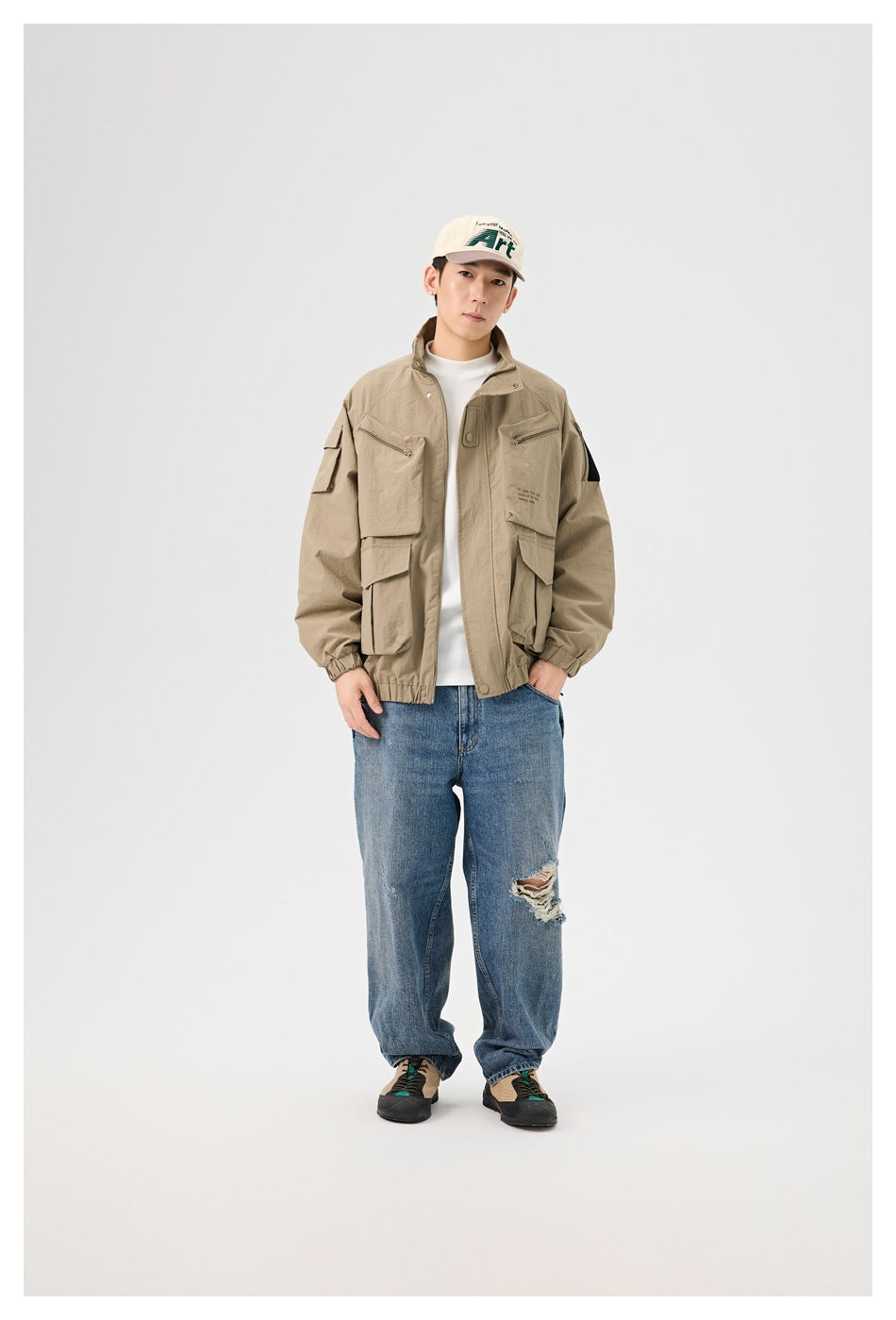 Man wearing a khaki utility jacket over a white top with blue ripped jeans, standing full length
