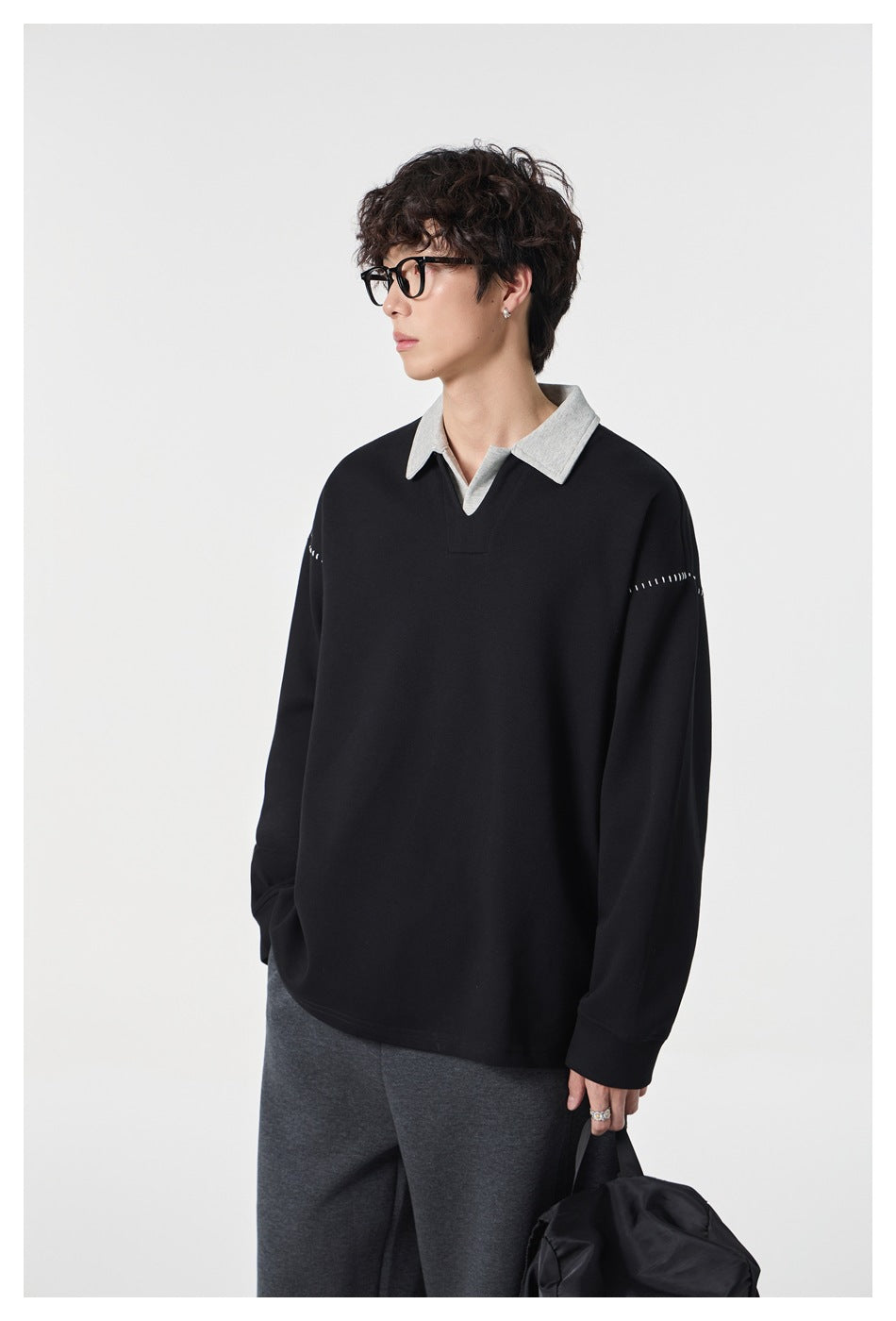 Model wearing the black pullover with gray polo collar and white stitching, gray trousers, holding a black bag