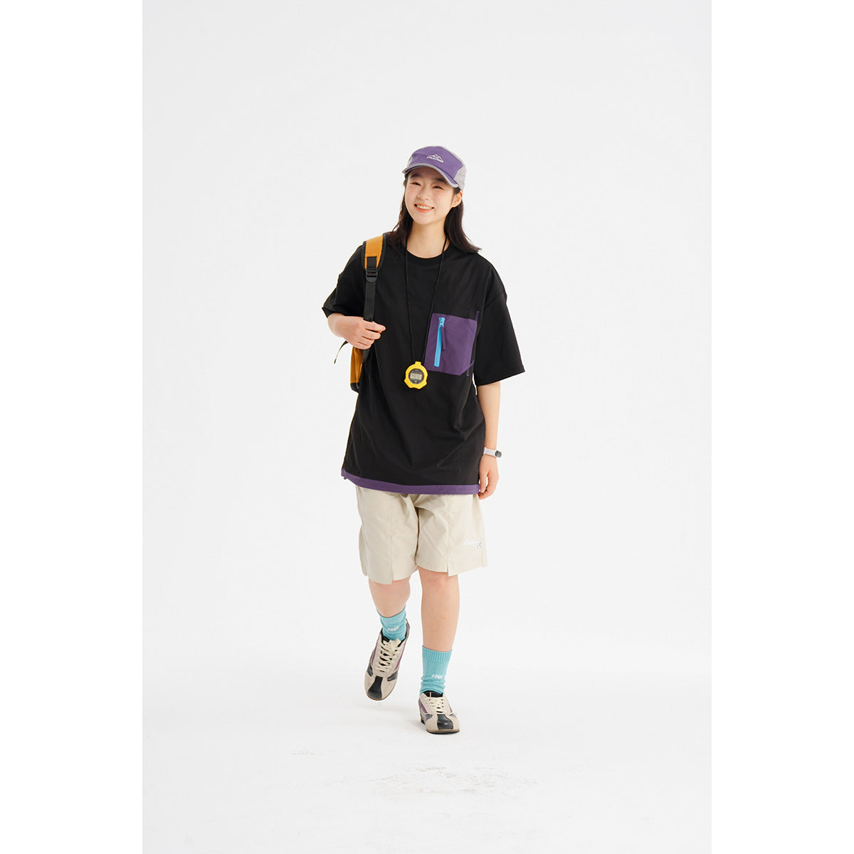 Model wearing a black oversized T-shirt with a purple zip chest pocket, beige shorts, and a purple cap.
