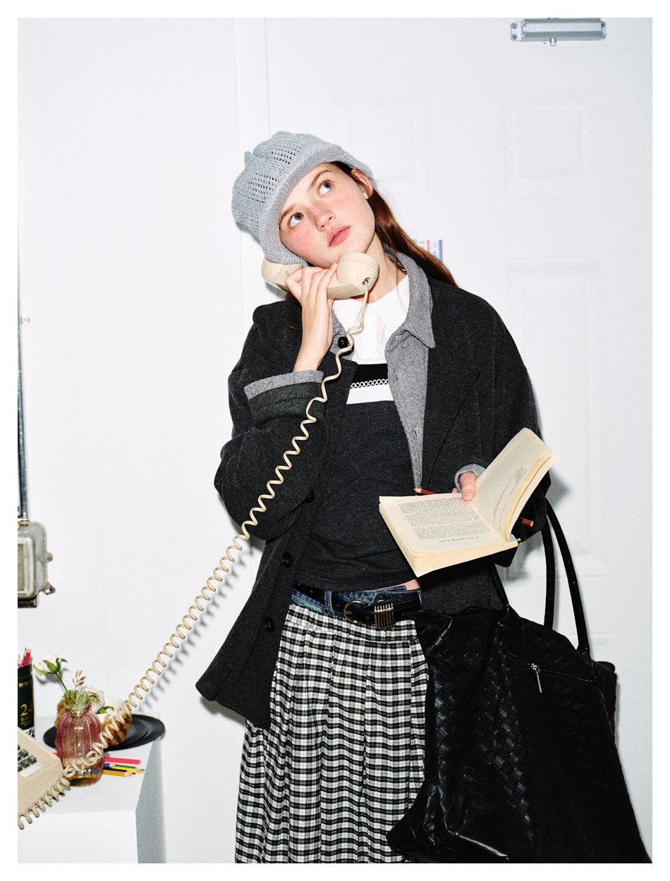 Model wears a charcoal jacket with contrasting gray collar and cuffs, a dark top and checked skirt while holding a book.