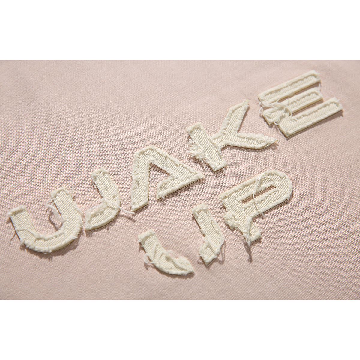 Close-up of cream raised “WAKE UP” lettering stitched onto pale blush-pink T-shirt fabric.
