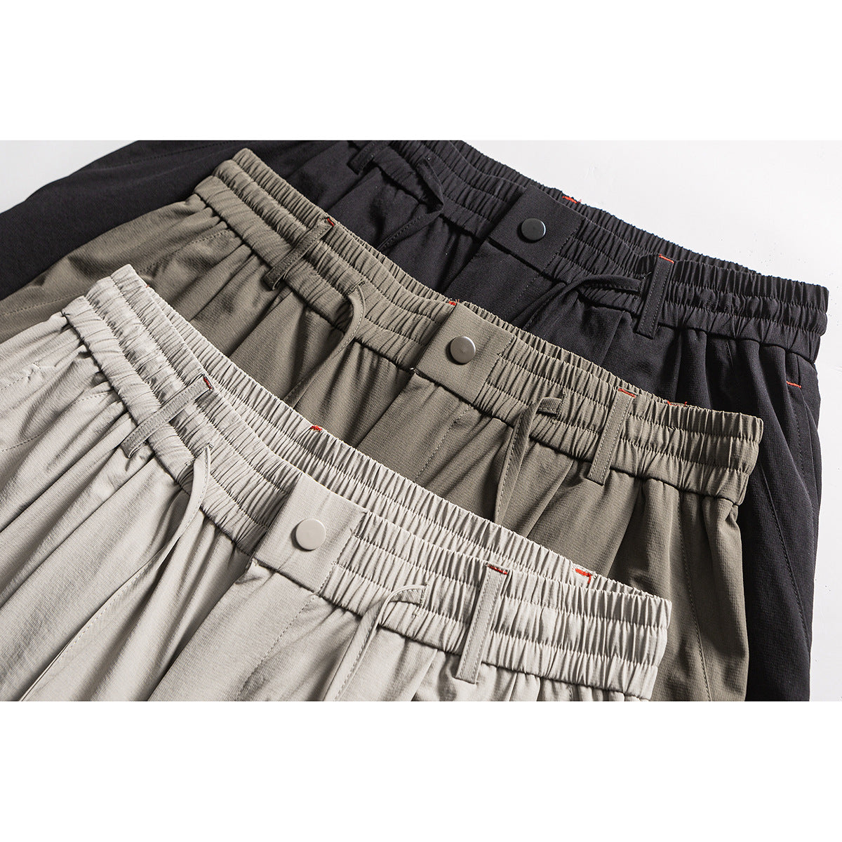 Close-up of three pairs of trousers in light gray, olive gray, and black, showing elastic waistbands and snap closures.