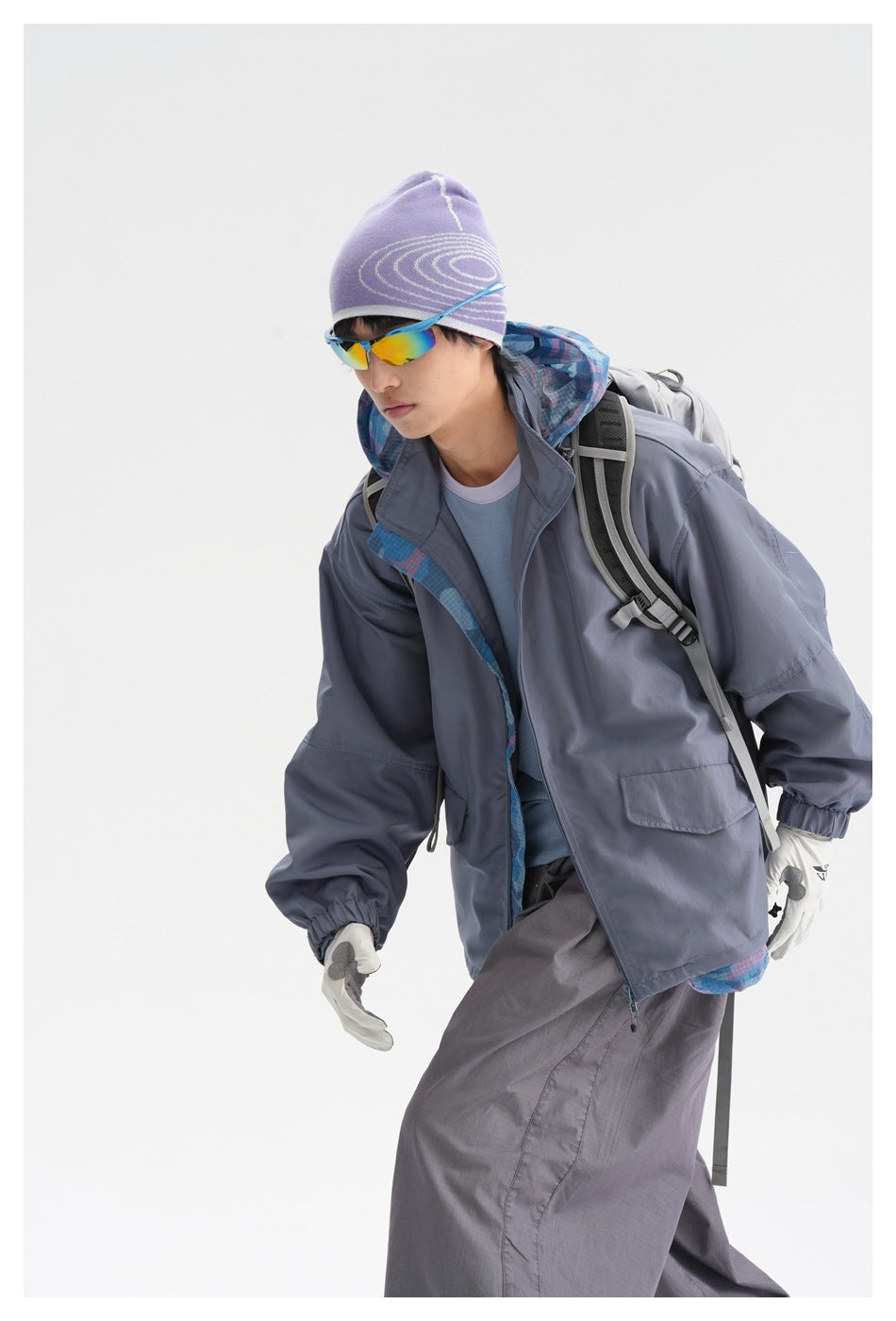 Model wearing a slate gray hooded jacket with blue patterned lining, loose gray pants, a lavender beanie and sunglasses.
