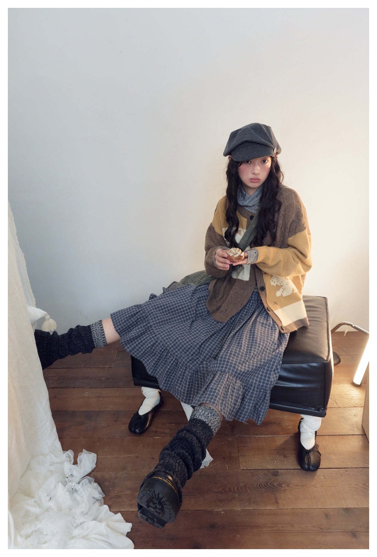 Model seated in a brown and mustard color-block cardigan, blue plaid skirt and gray cap.