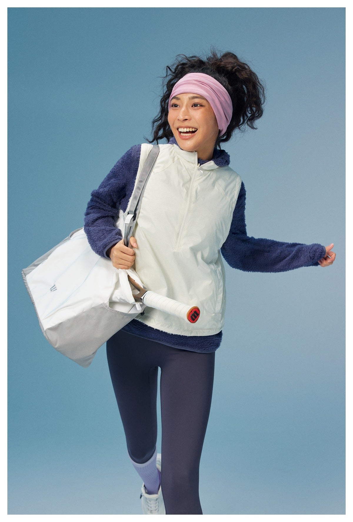 Model wearing an off-white zip vest over a navy fleece top and dark leggings, carrying a large light-colored bag.